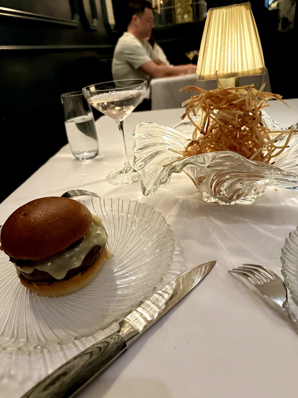 Le Burger at Le B. on a ribbed glass plate, served alongside a tall glass bowl of ultra-thin crispy shoestring potatoes, with a martini in the background